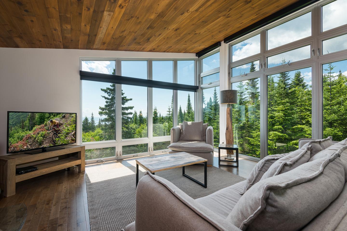 Salon moderne avec grandes baies vitrées donnant sur une forêt, canapé beige et TV allumée.