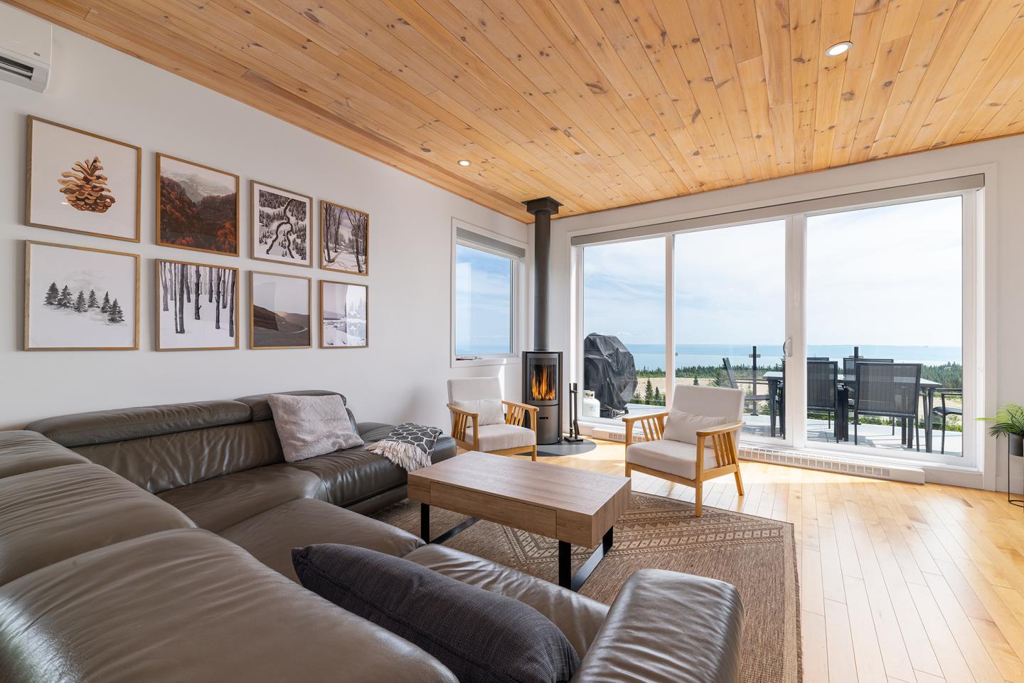 Salon moderne avec canapé en cuir, poêle, grandes fenêtres surplombant la mer.