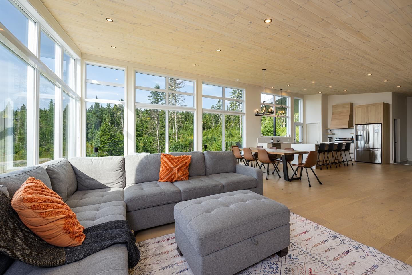 Salon moderne avec canapé gris, coussins orange, grandes fenêtres, vue sur forêt.