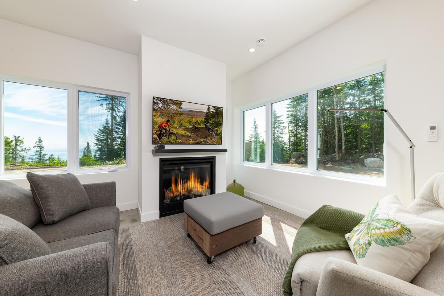 Salon lumineux avec cheminée, canapés gris et grande fenêtre donnant sur une forêt.
