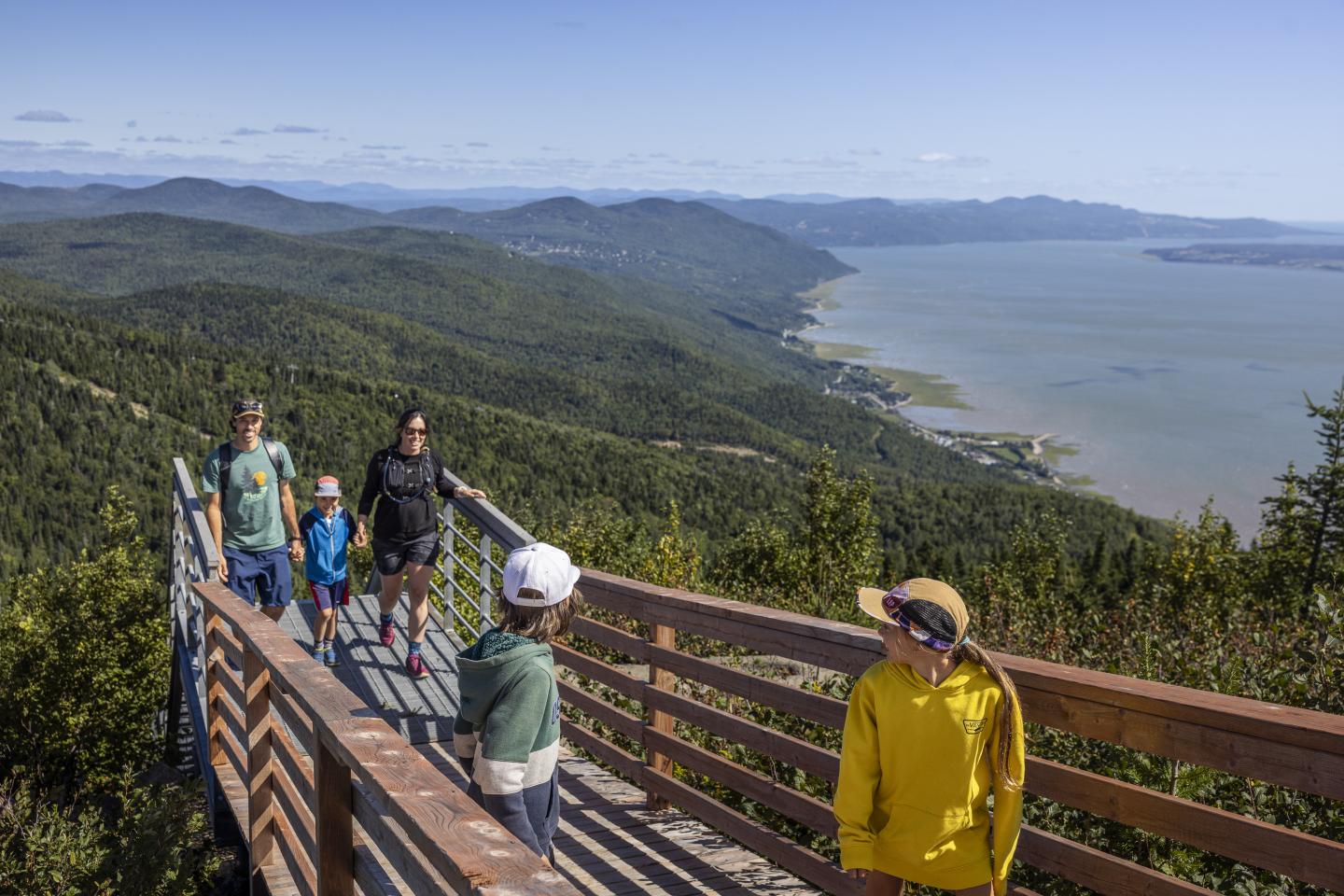 Famille se promenant sur une passerelle en bois avec vue sur la montagne et la mer.