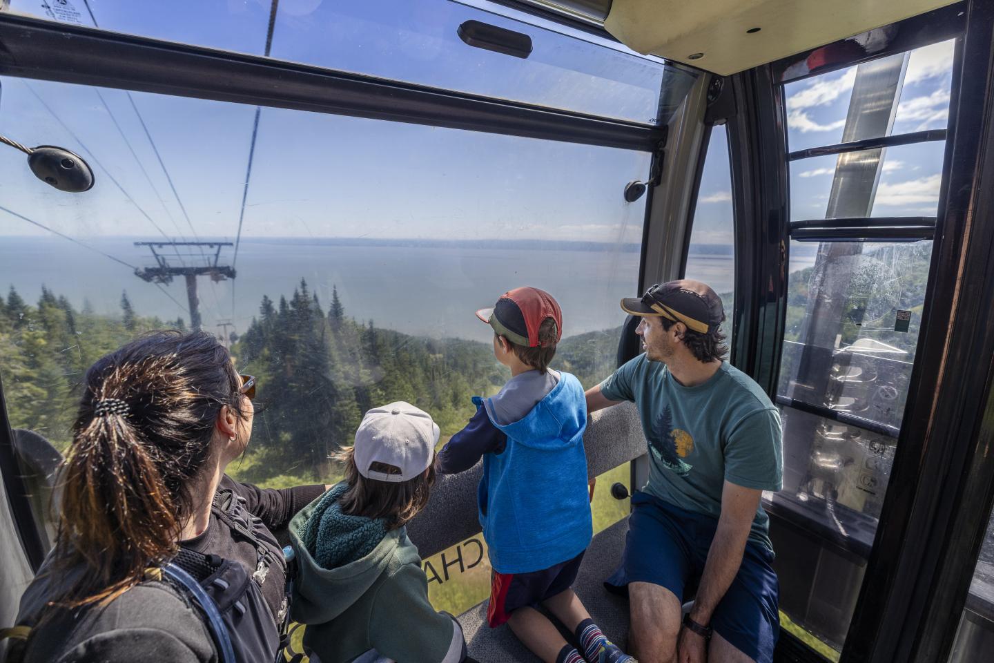 Quatre personnes dans une télécabine, vue sur la mer et forêt en contrebas.