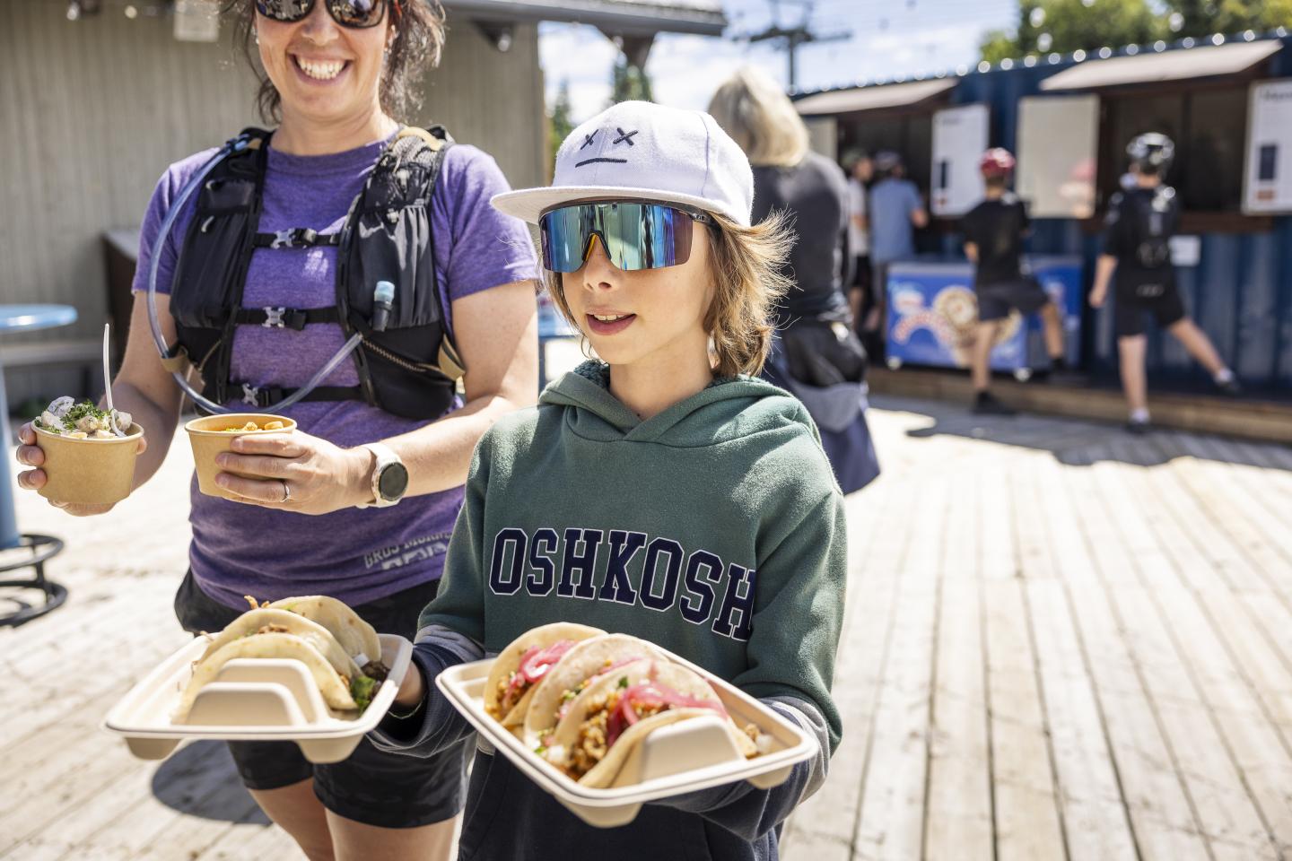 Enfant et adulte souriant portant des plateaux de tacos en extérieur.