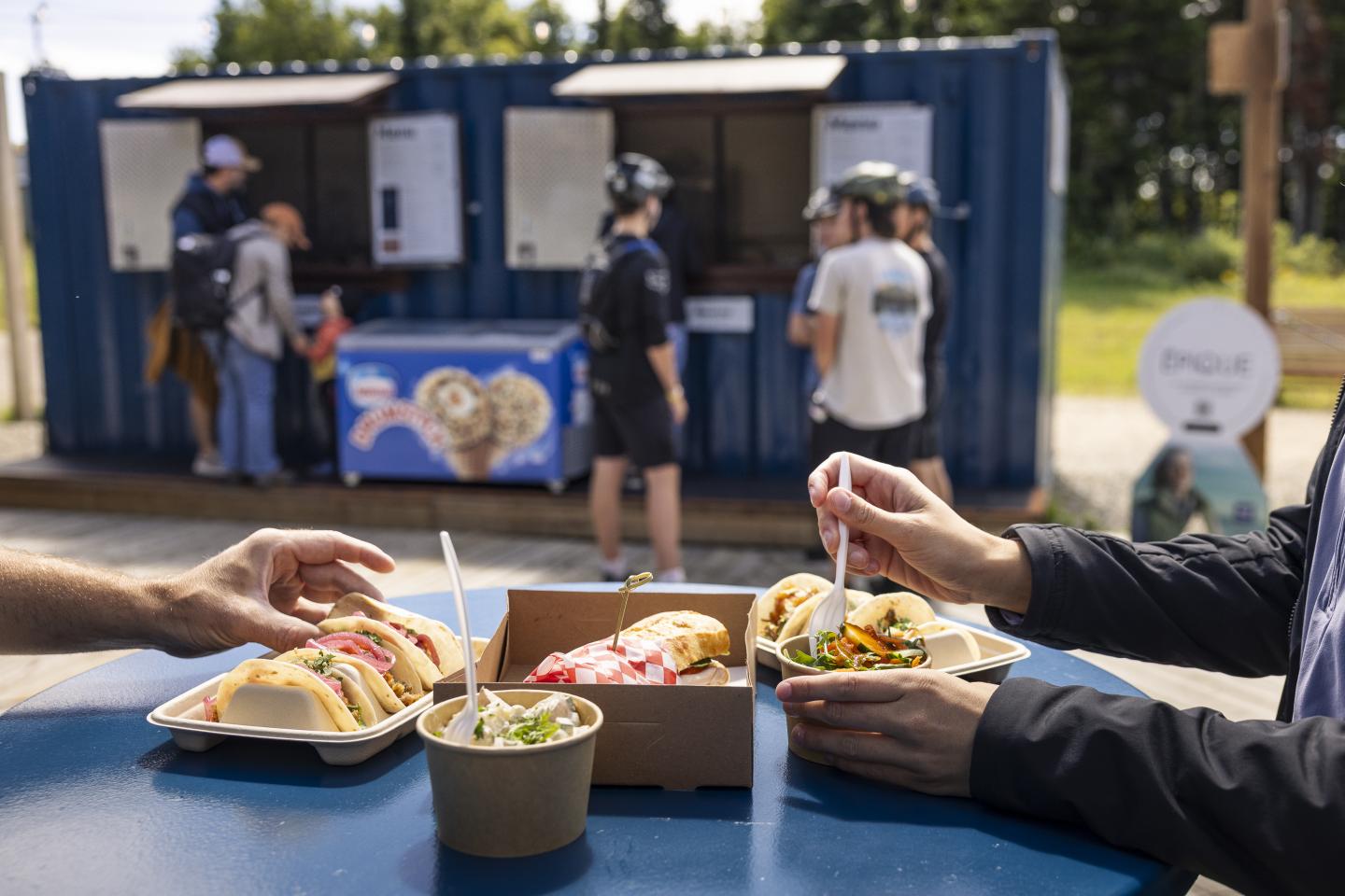 Des personnes dégustent des plats à une table extérieure près d'un stand de nourriture.