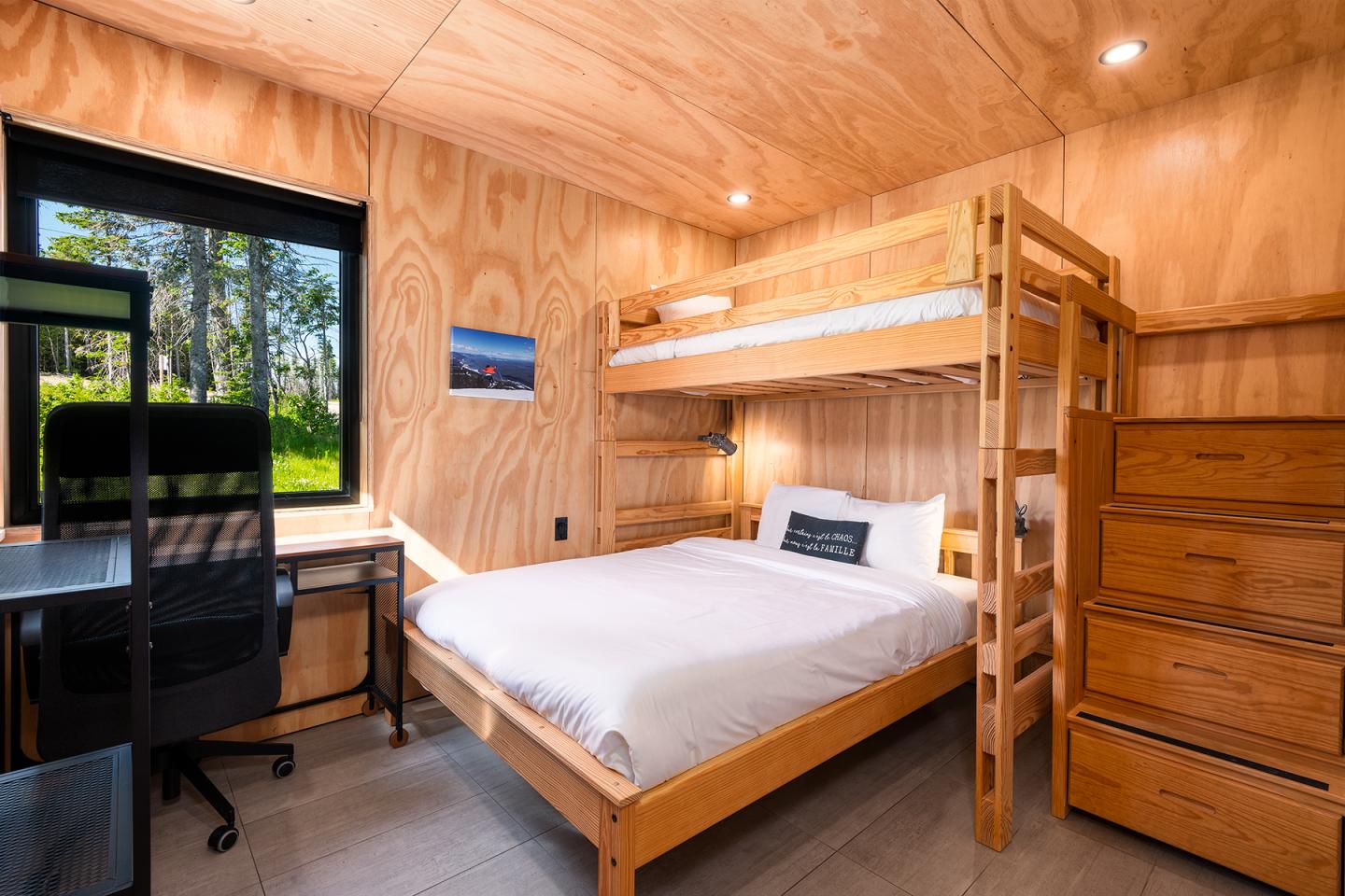 Petite chambre en bois avec lit superposé, bureau et commode, fenêtre sur jardin.