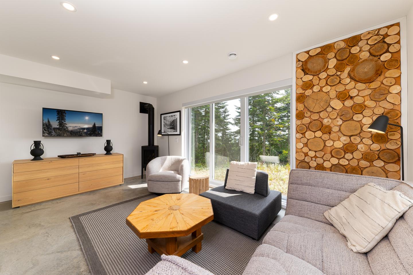 Salon moderne avec canapé gris, table basse en bois, grande fenêtre, mur décoratif en rondins.