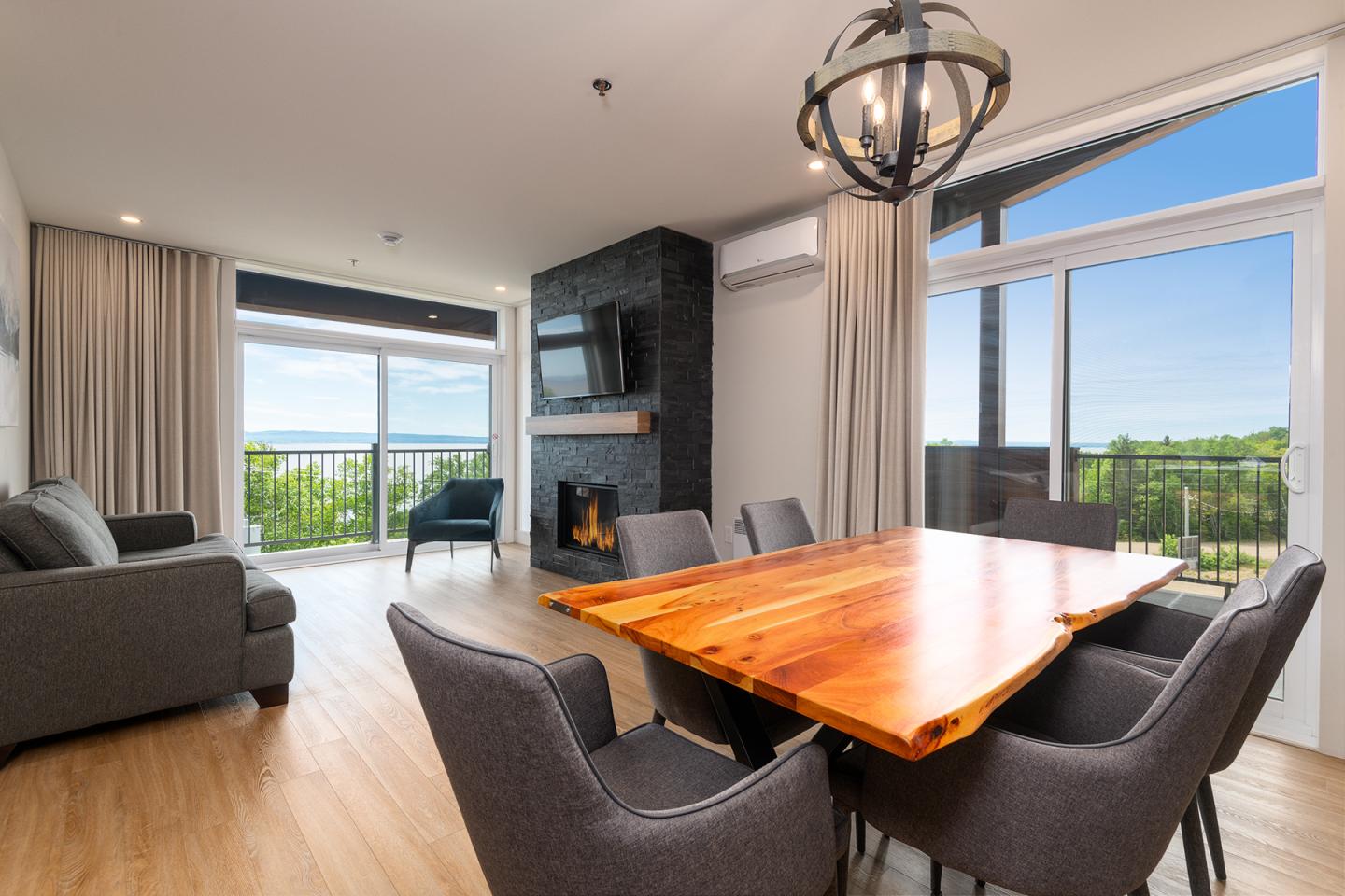 Salon moderne avec table en bois, canapés gris, cheminée noire et vue sur jardin.