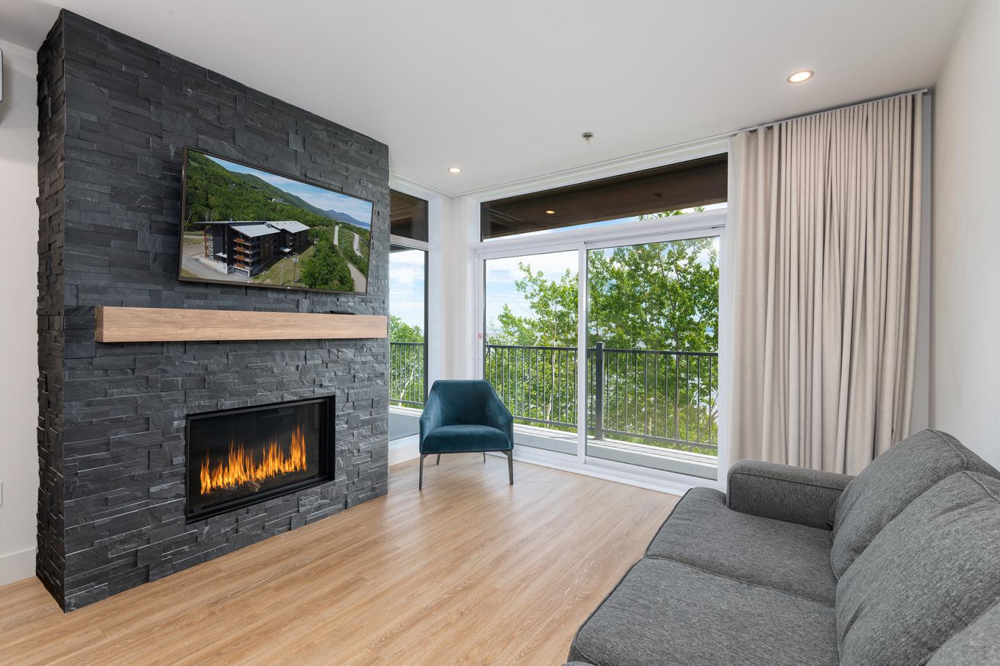 Salon moderne avec cheminée, fauteuil bleu, canapé gris, grande fenêtre avec vue sur la verdure.