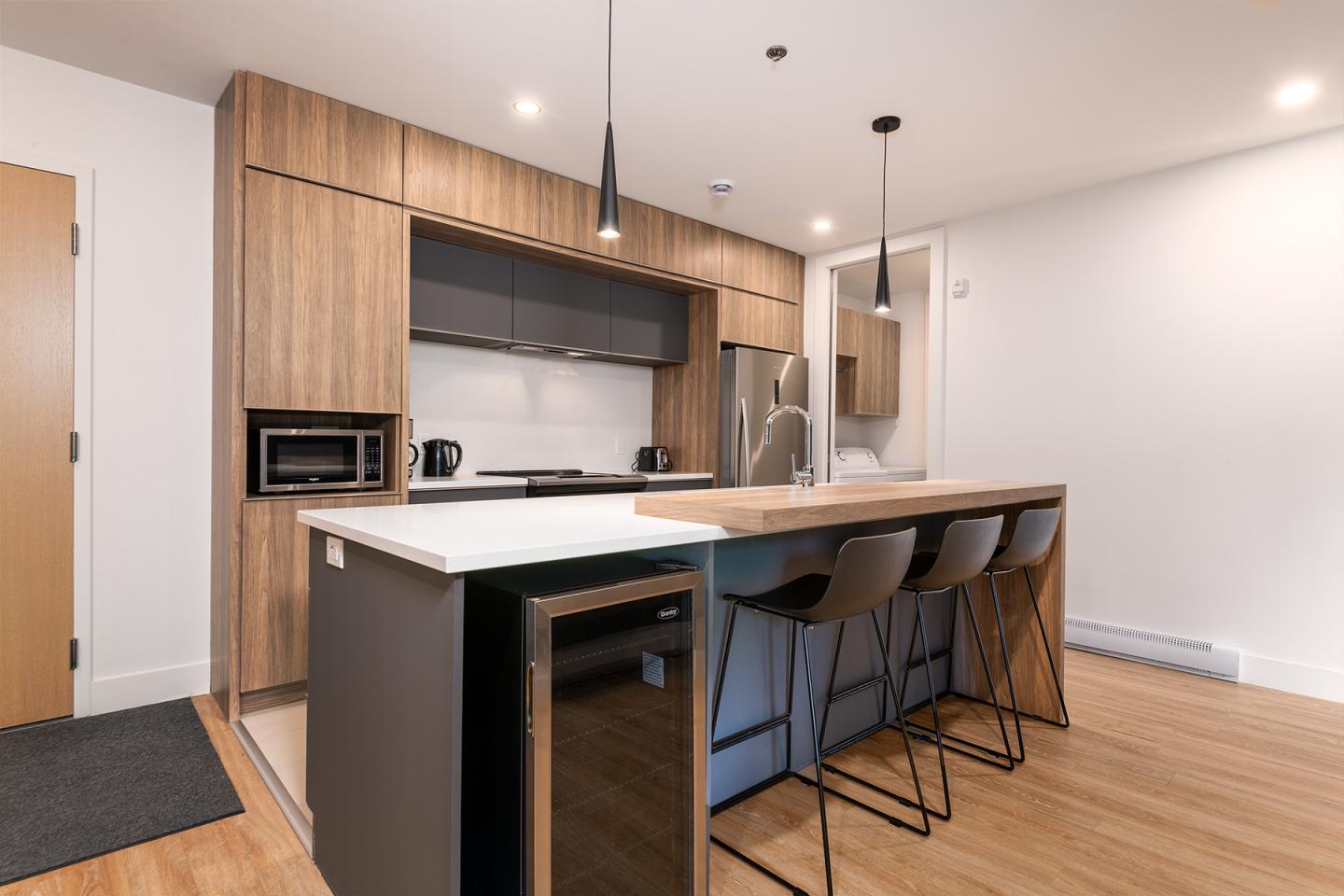 Cuisine moderne avec îlot central, bois clair, chaises noires, et éclairage minimaliste.