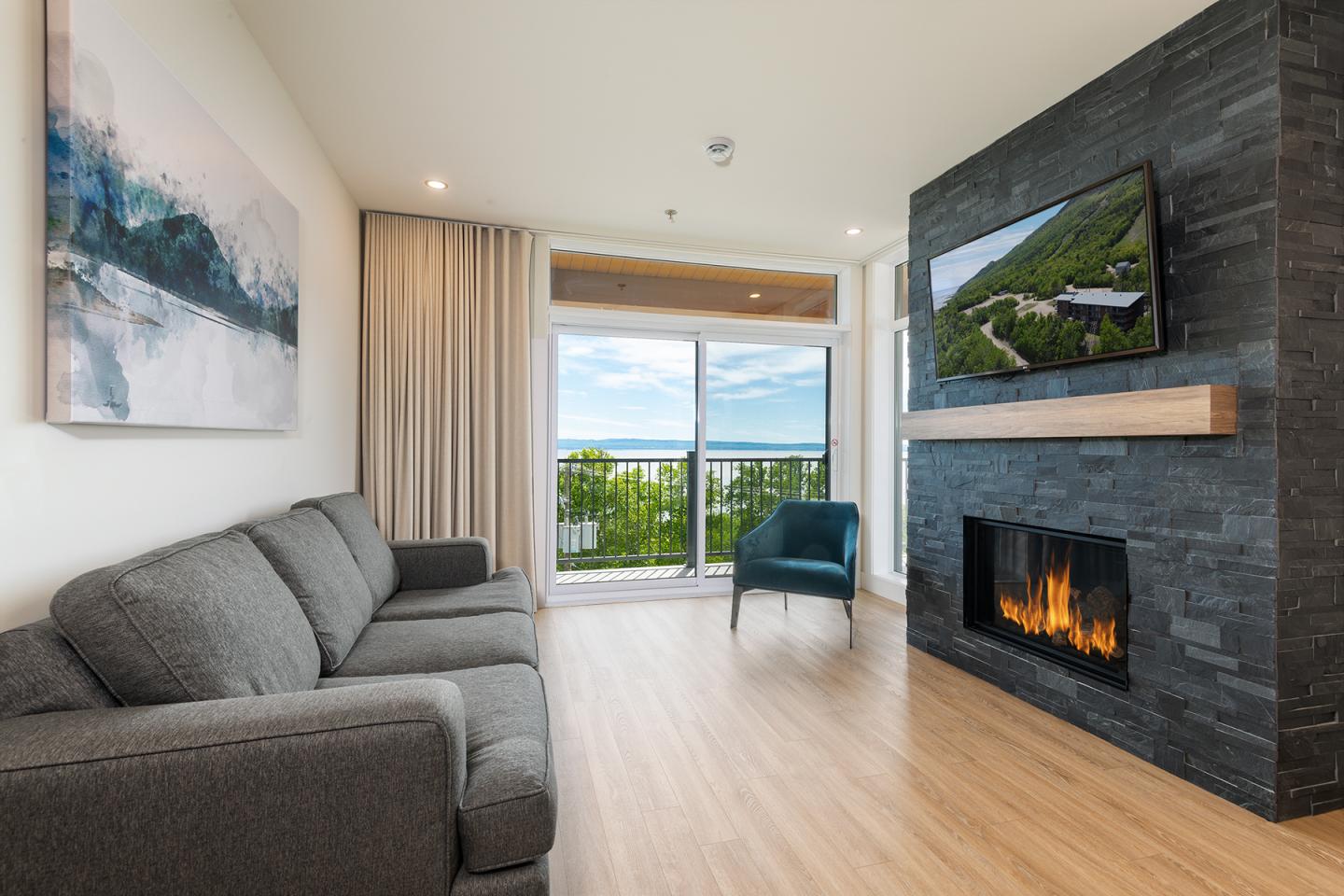 Salon moderne avec cheminée, canapé gris, fauteuil bleu, vue sur la mer par la fenêtre.