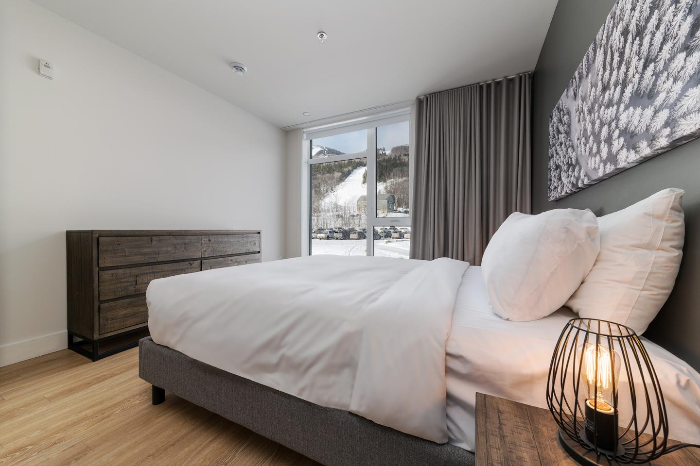 Chambre moderne avec lit, commode, lampe allumée et grande fenêtre.