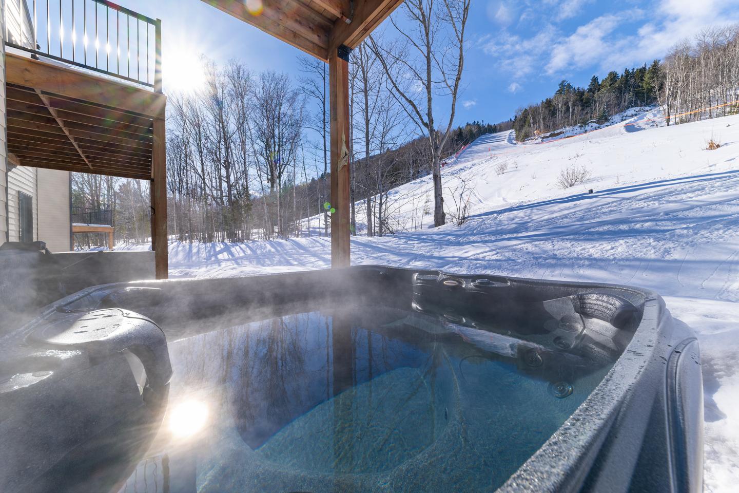 Jacuzzi fumante sous un balcon, paysage enneigé en arrière-plan.