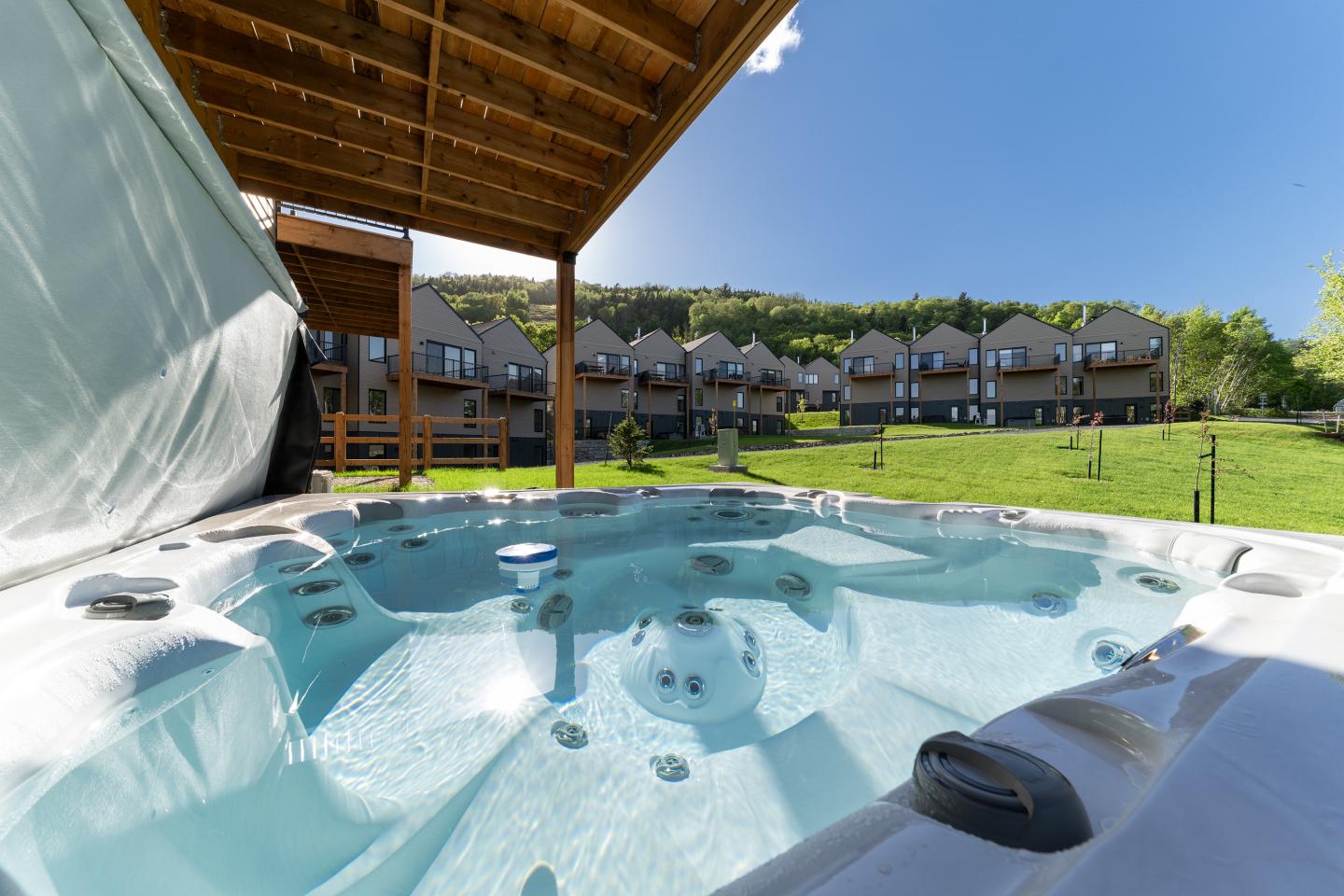 Jacuzzi extérieur sous abri, vue sur pelouse et maisons, ciel bleu.
