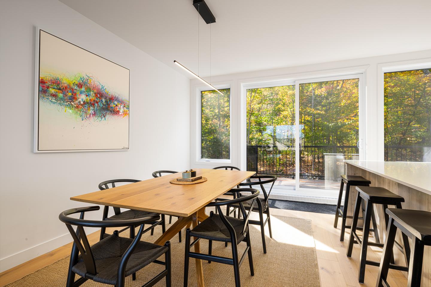 Salle à manger lumineuse avec table en bois, tableau coloré et grandes fenêtres.