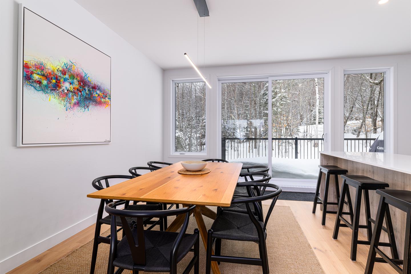 Salle à manger moderne, grande table en bois, chaises noires, tableau coloré au mur.