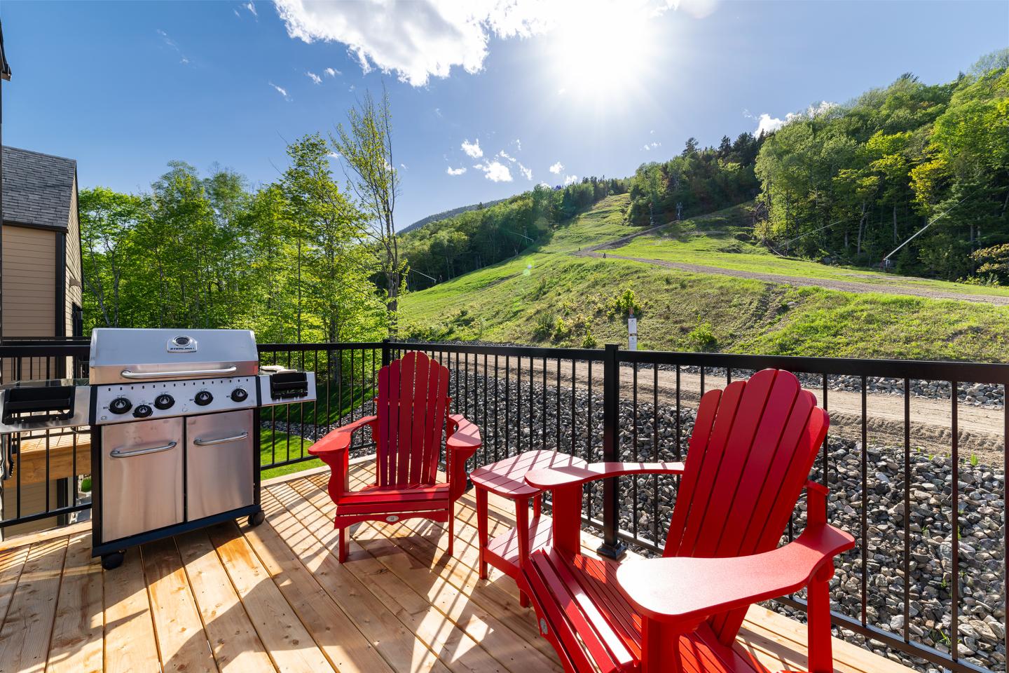 Deux chaises rouges sur une terrasse ensoleillée avec un barbecue, vue sur colline verdoyante.
