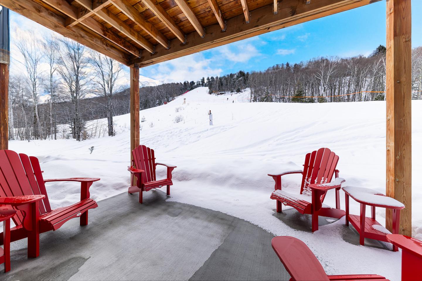 Chaises rouges sous une terrasse en bois devant un paysage enneigé.