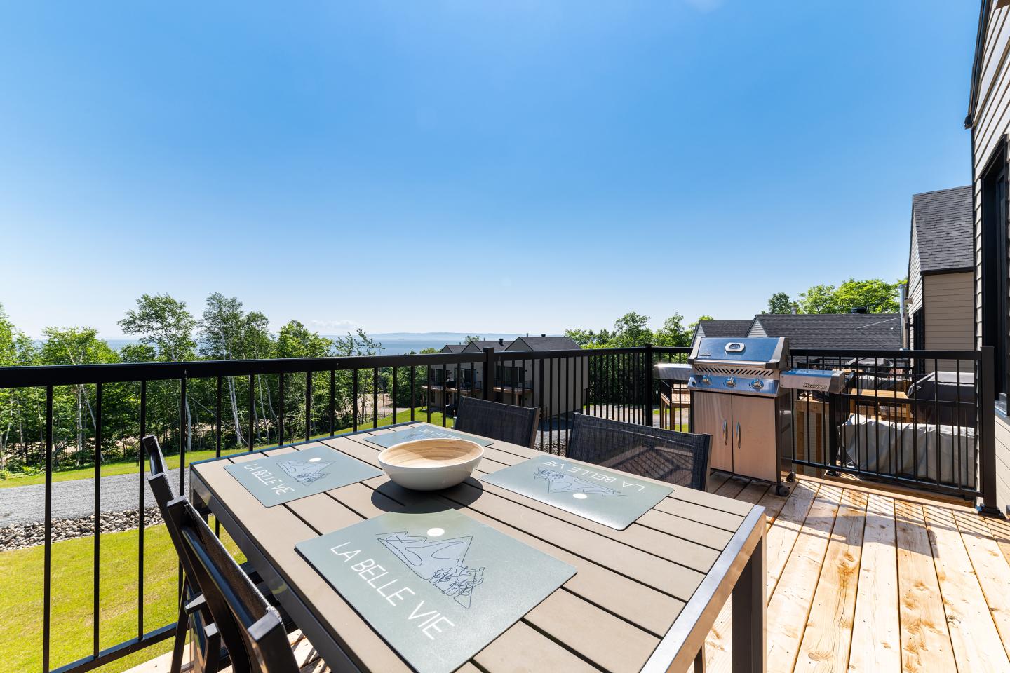 Terrasse en bois avec table dressée, barbecue et vue sur le jardin sous un ciel bleu.