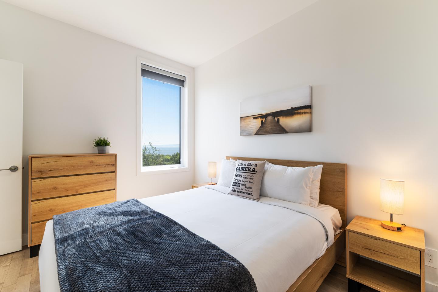Chambre lumineuse avec lit double, commode en bois, fenêtre et tableau au mur.