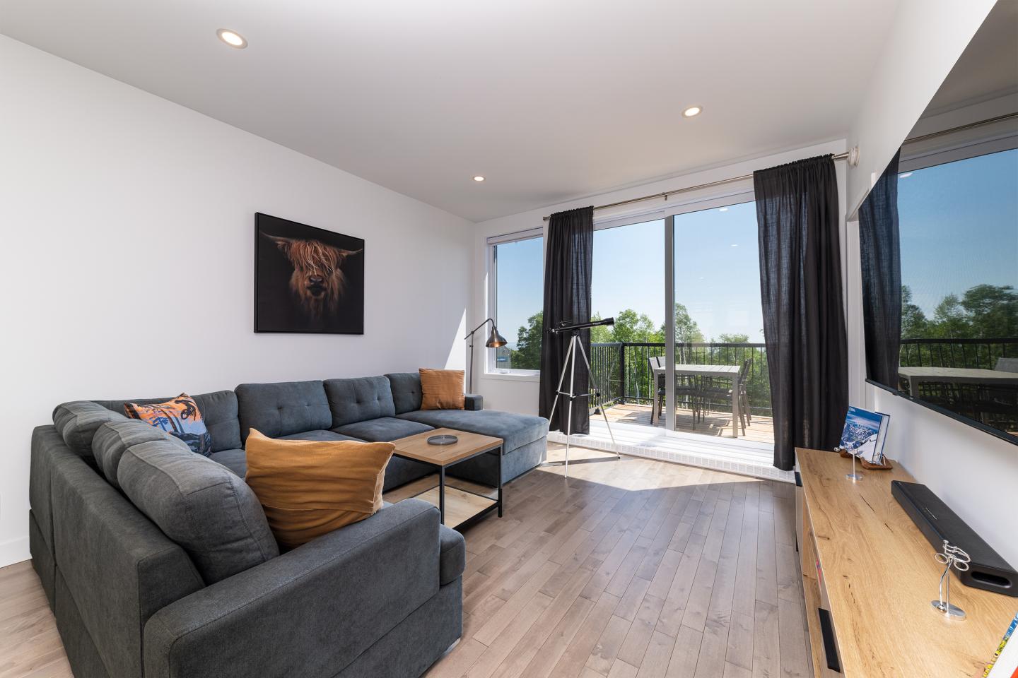 Salon moderne avec canapé gris, coussins orange, grandes fenêtres et balcon ensoleillé.