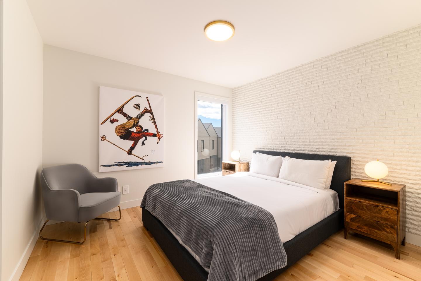 Chambre moderne avec lit double, chaise grise, tableau et lumière douce.