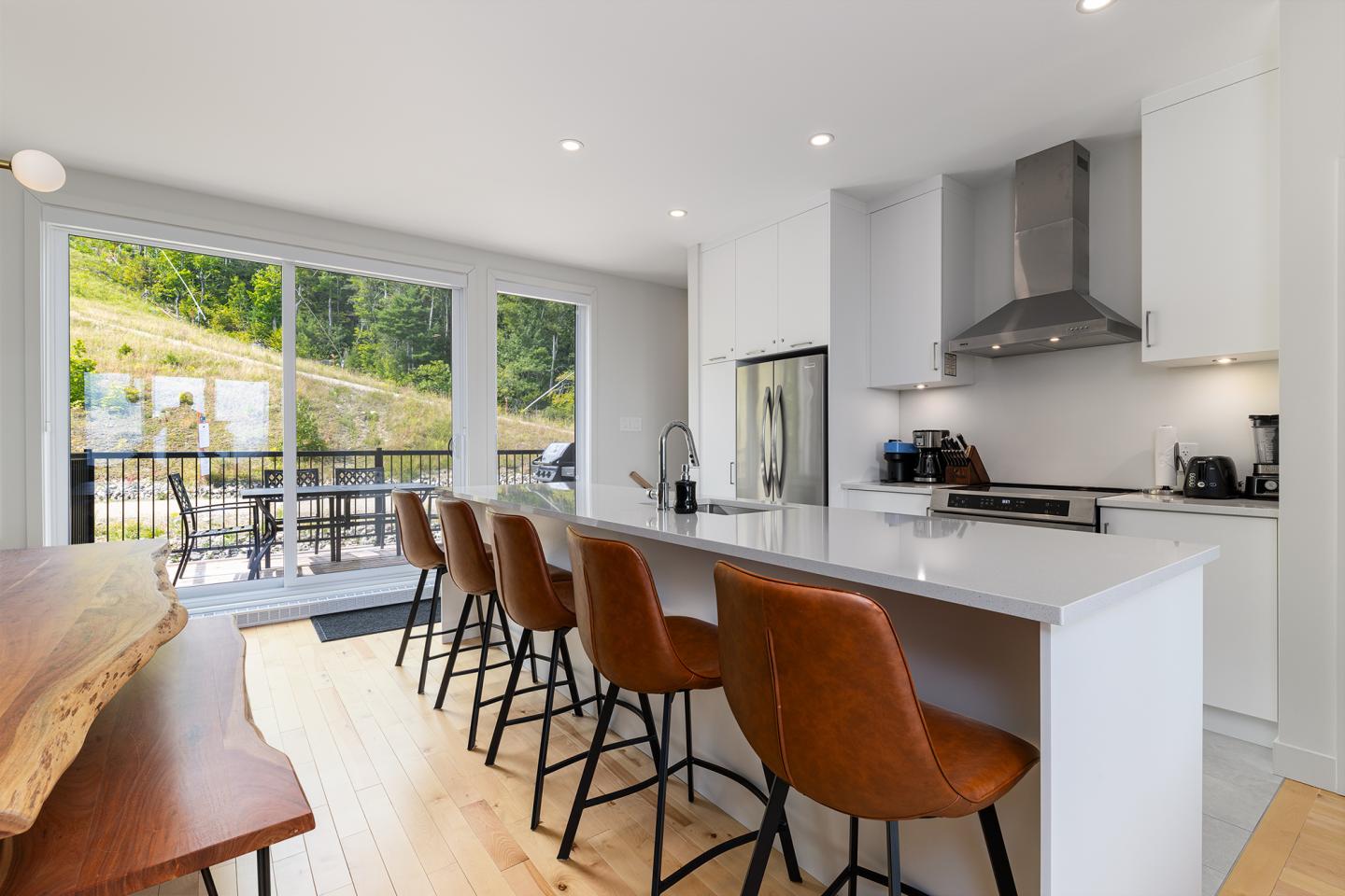 Cuisine moderne avec îlot central, chaises marron, et grande baie vitrée.