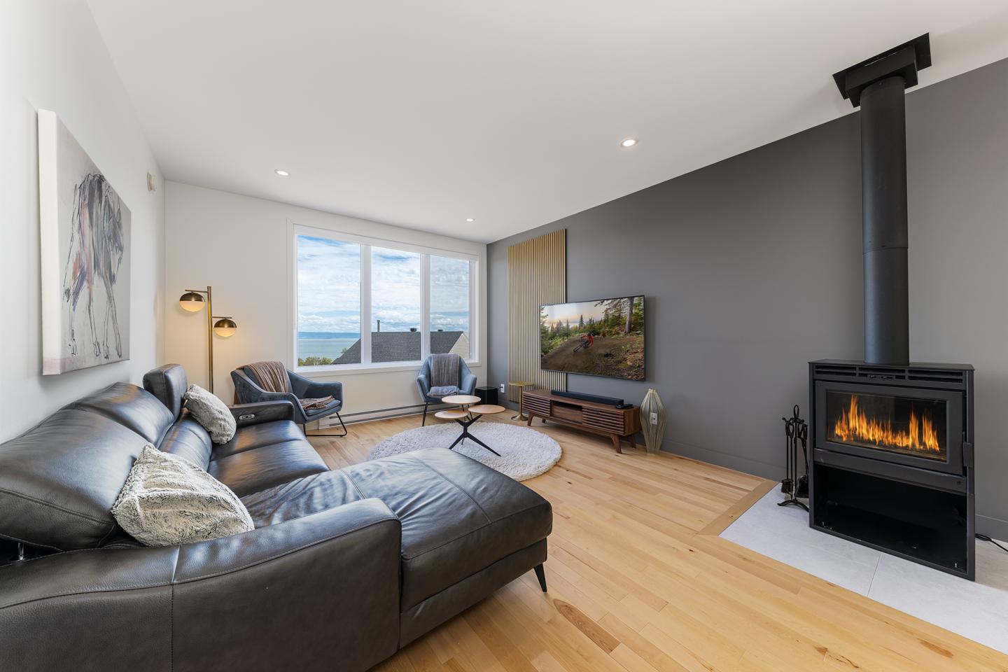 Salon moderne avec canapé en cuir, poêle à bois et vue sur la mer.