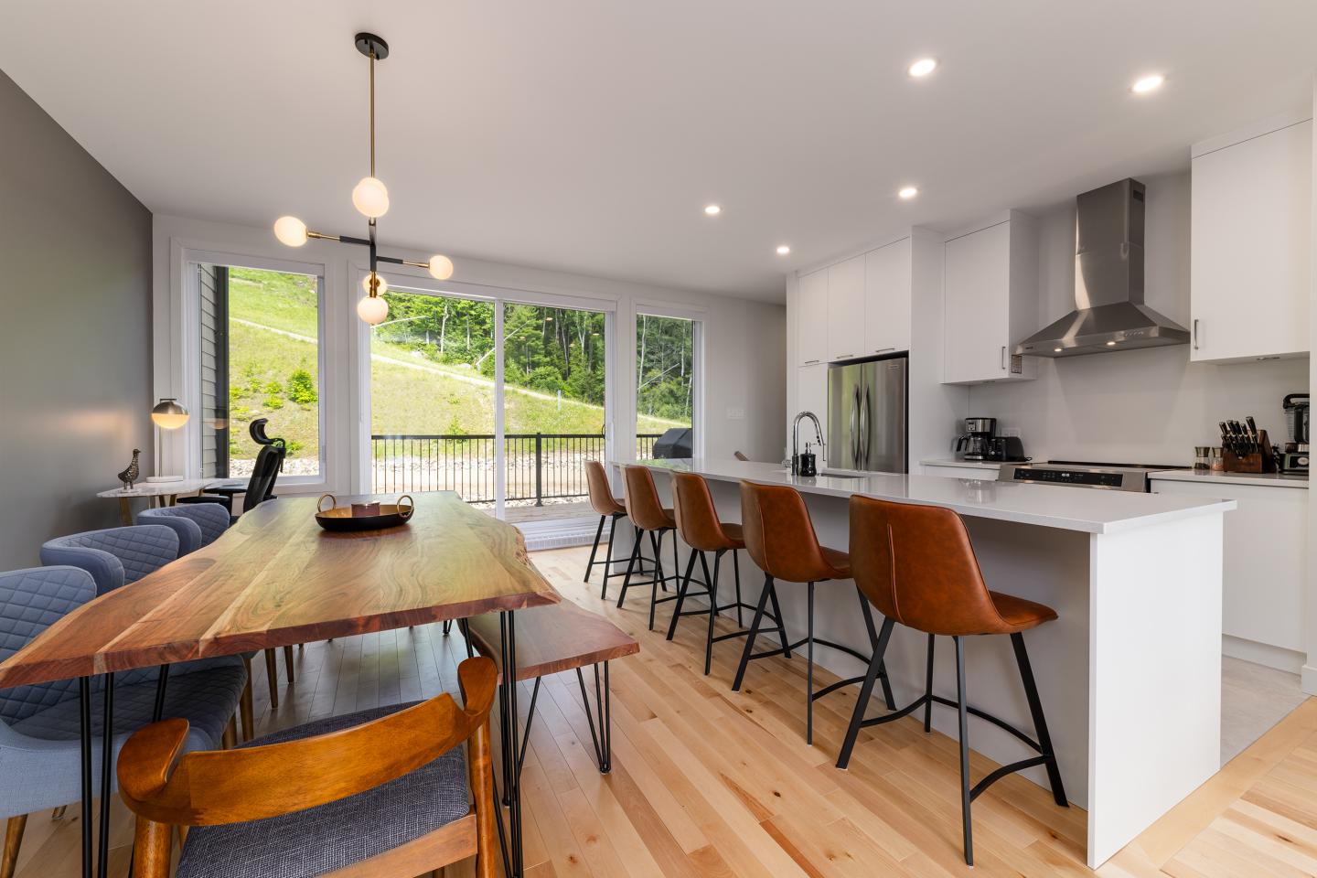 Cuisine moderne avec îlot central, chaises hautes brunes et grande table en bois.
