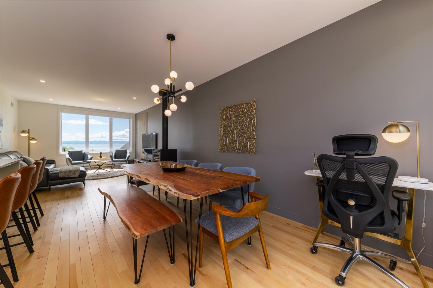 Salle à manger moderne avec table en bois, chaises, mur gris et éclairage suspendu.