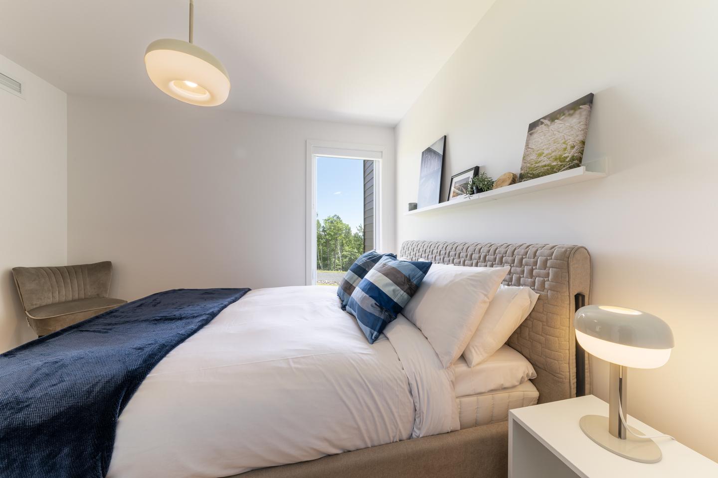 Chambre moderne avec lit, coussins bleus, lampe et fenêtre ensoleillée.