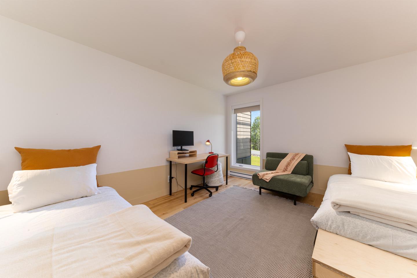 Chambre lumineuse avec deux lits, bureau et fauteuil vert.