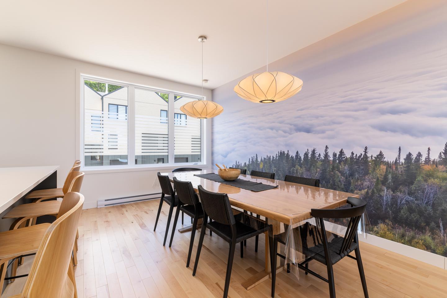 Salle à manger moderne avec grande table en bois, chaises noires et mural de paysage forestier.