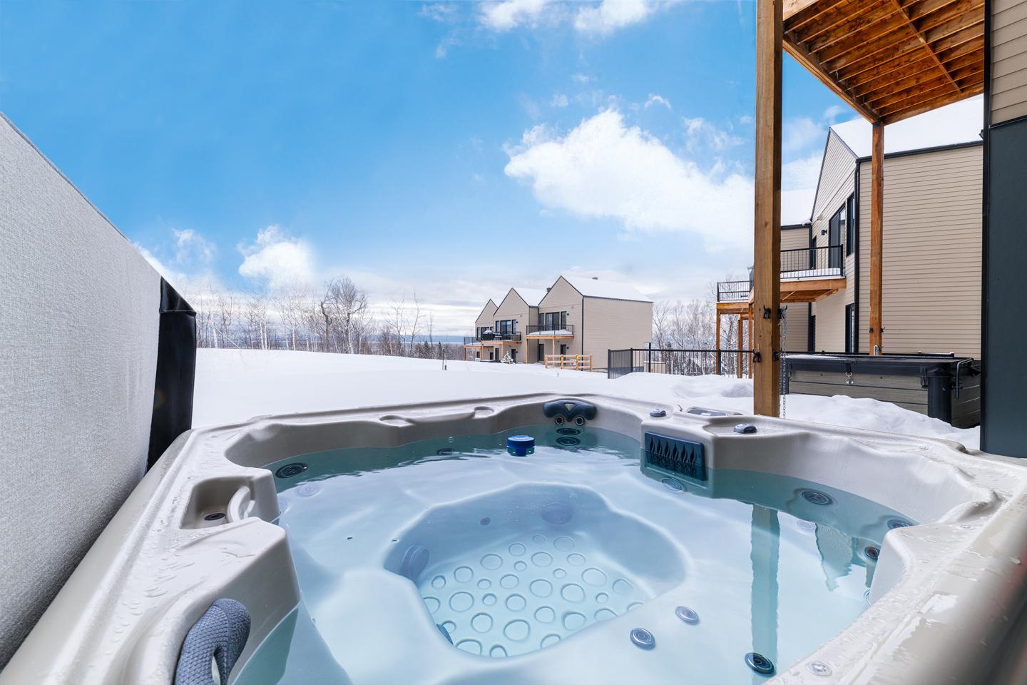 Bain à remous extérieur enneigé avec vue sur des maisons et ciel bleu.
