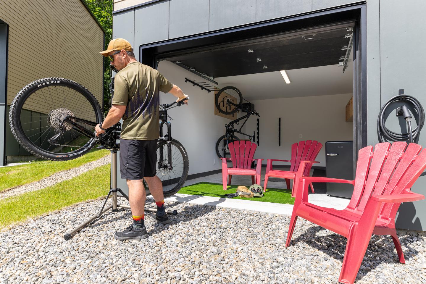 Homme réparant un vélo devant un garage avec deux chaises roses.