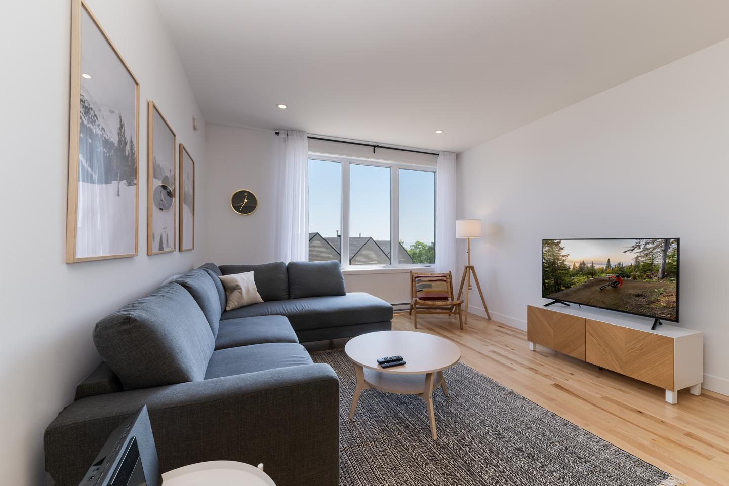 Salon moderne avec canapé gris, téléviseur et grandes fenêtres.