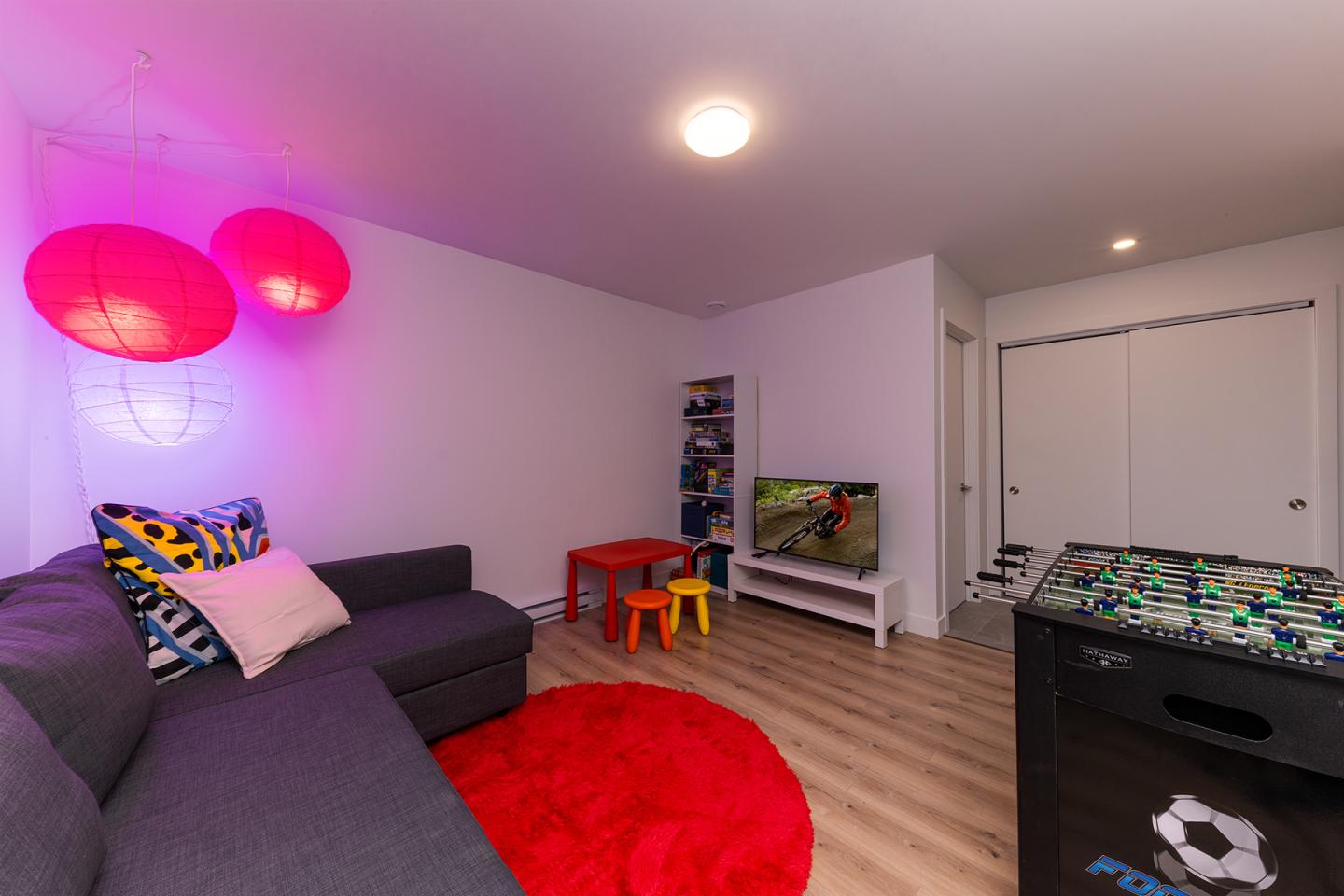 Salle de jeux avec canapé, table rouge, lampe rose et baby-foot.