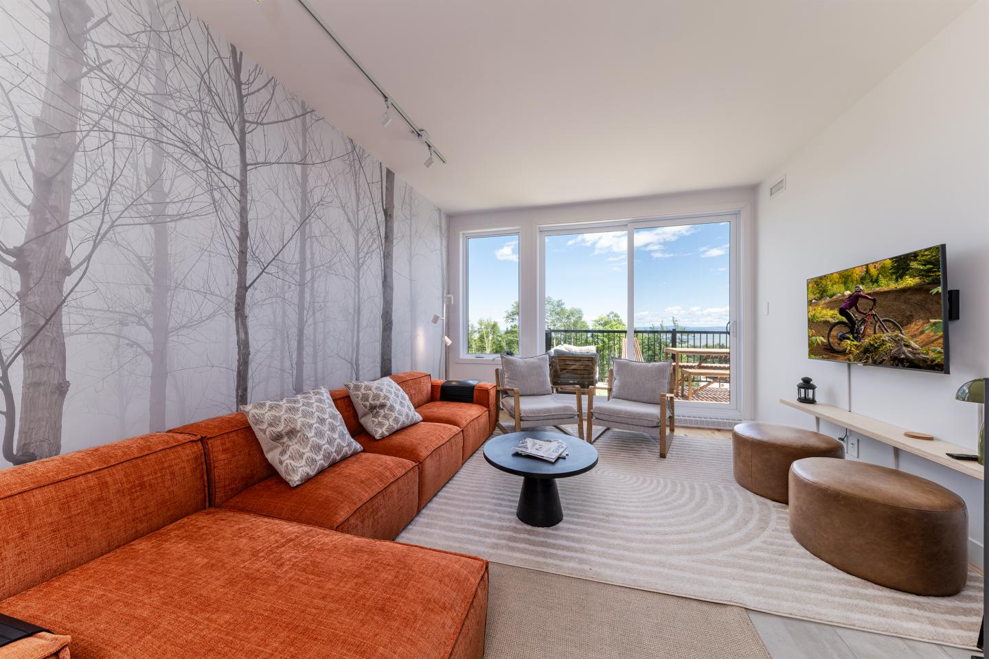 Salon moderne avec canapé orange, papier peint arbre, télé, et vue sur jardin par baie vitrée.