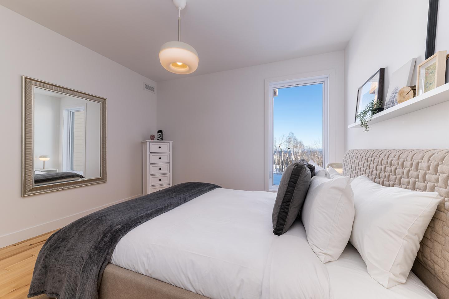 Chambre moderne avec lit blanc, grande fenêtre et miroir encadré.