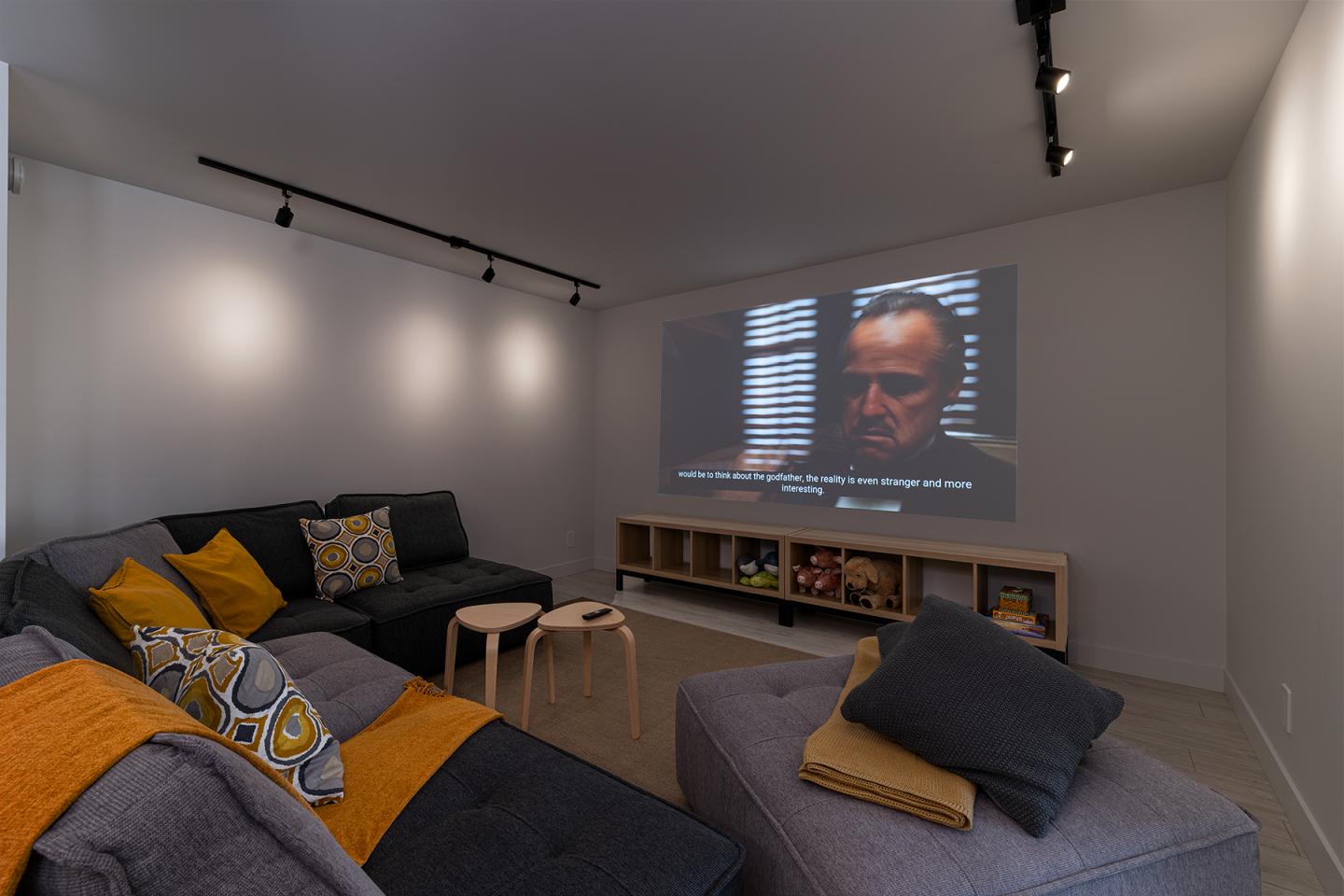 Salon moderne avec un canapé gris, coussins jaunes et un écran projetant un film.