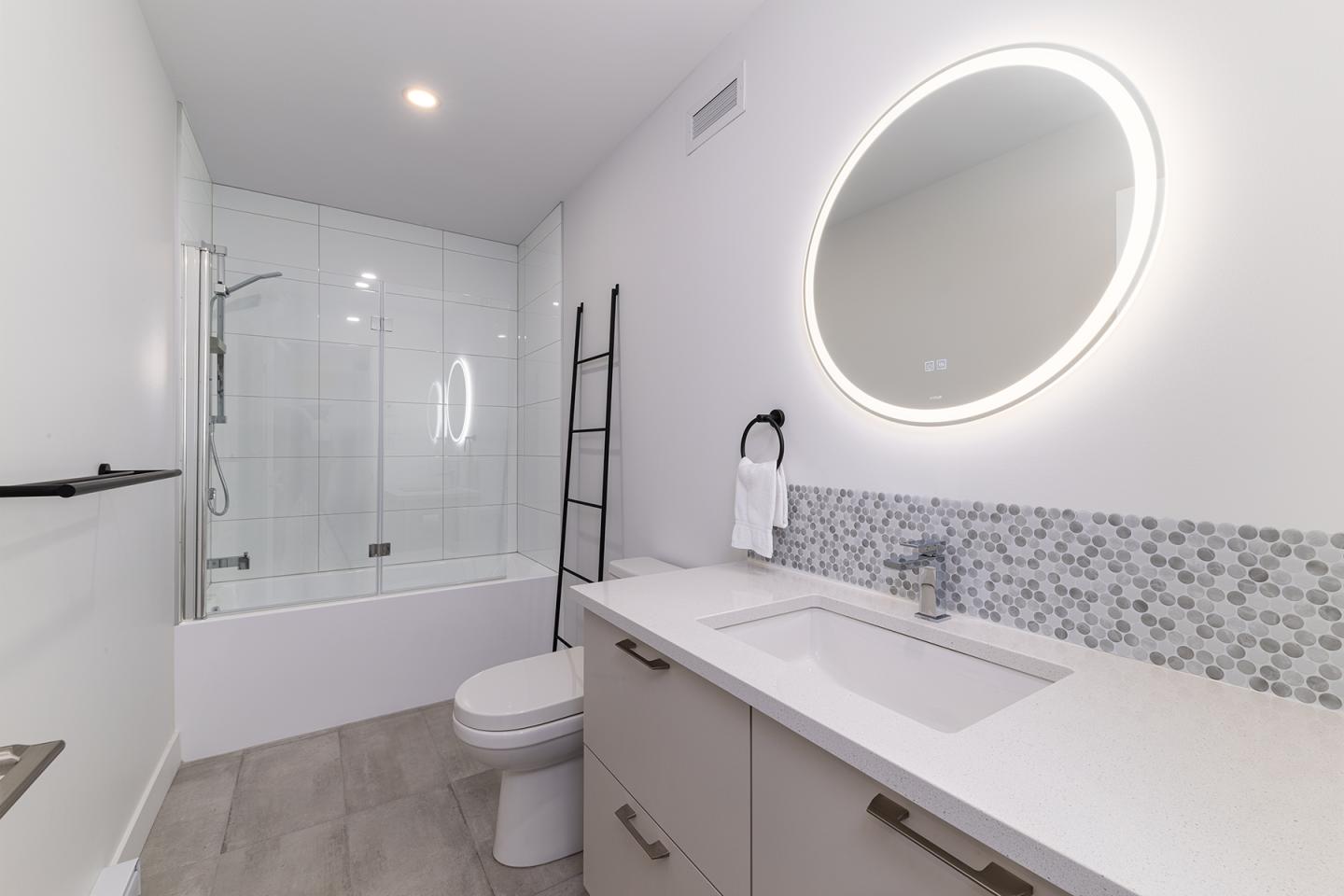 Salle de bain moderne, miroir rond éclairé, douche vitrée, échelle noire.