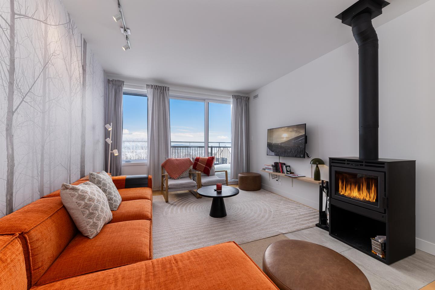 Salon moderne avec canapé orange, poêle à bois, TV et grande baie vitrée.