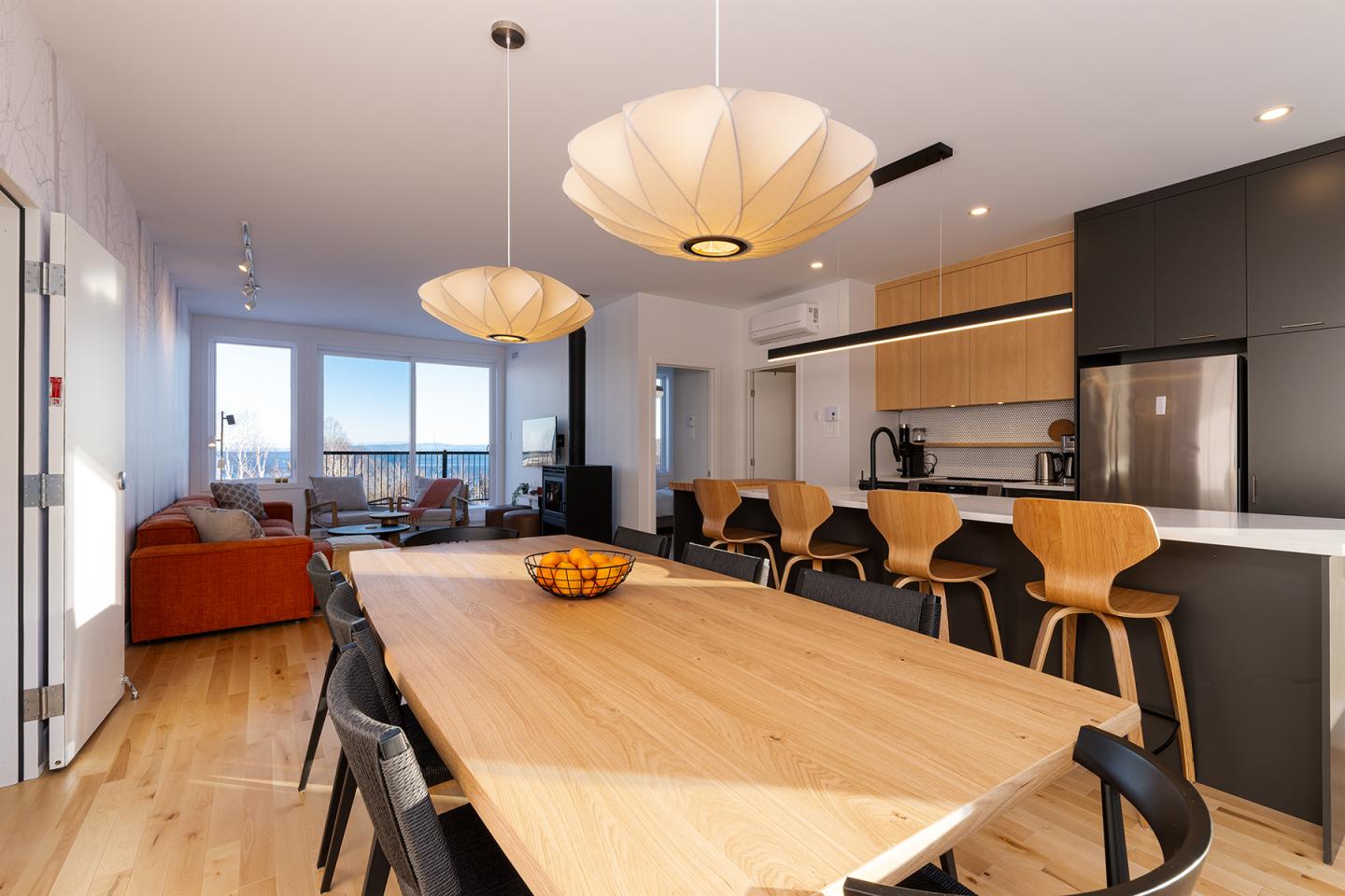 Salle à manger moderne avec table en bois clair, chaises noires, cuisine ouverte et canapé orange.