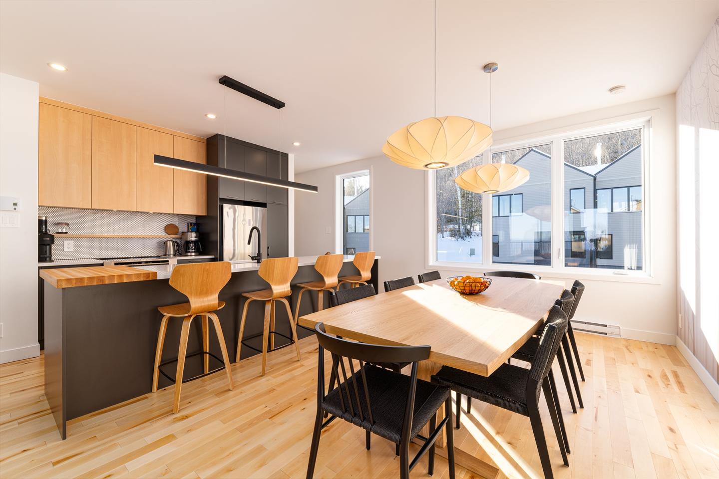 Cuisine moderne avec comptoir en bois, chaises hautes et table à manger éclairée par le soleil.