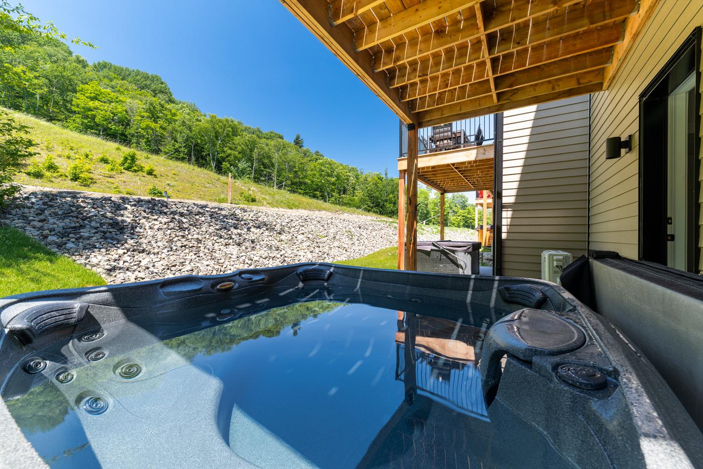 Jacuzzi extérieur sous terrasse en bois, ciel bleu et colline verdoyante.