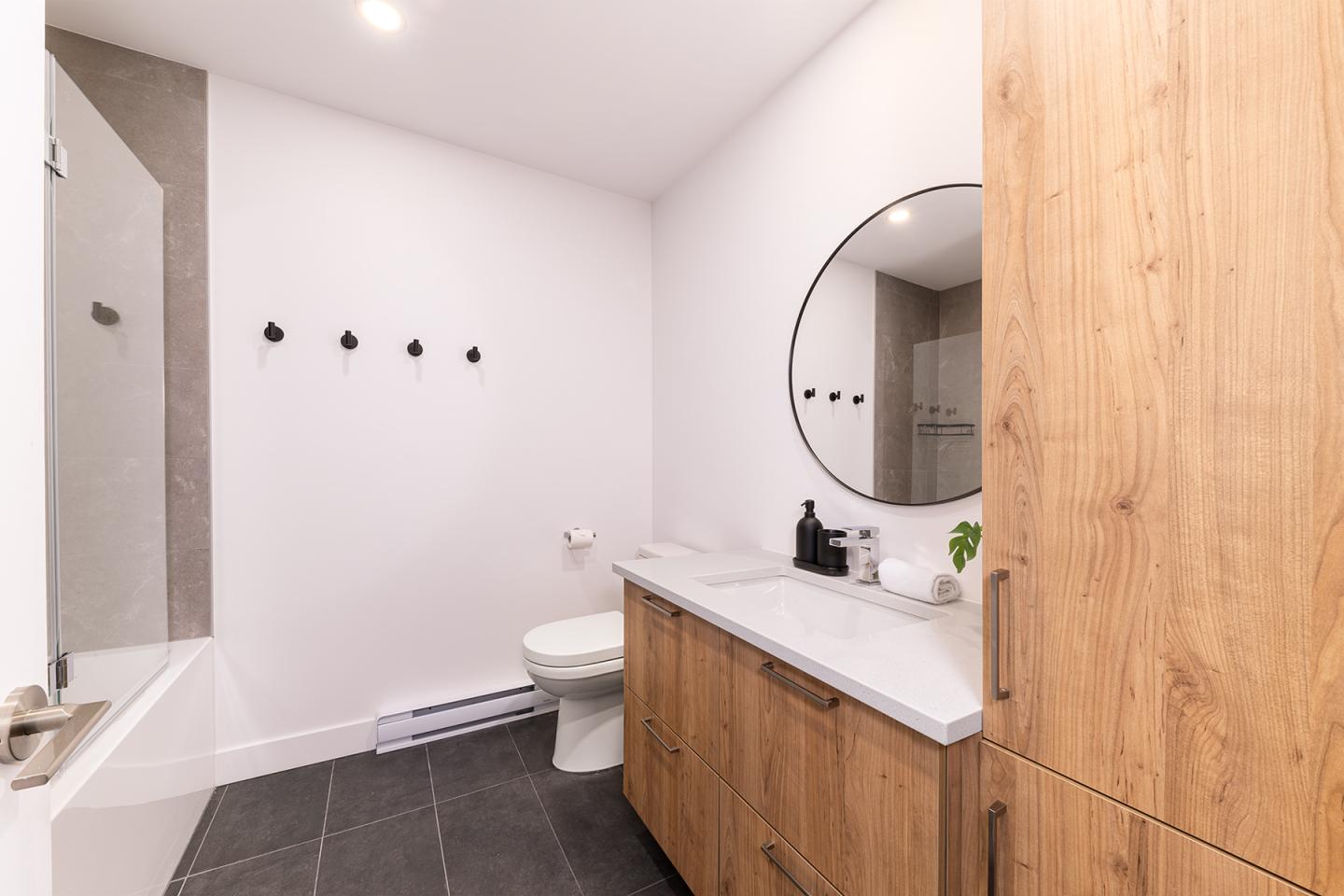 Salle de bain moderne avec mur blanc, meuble en bois clair, miroir rond et carrelage sombre.