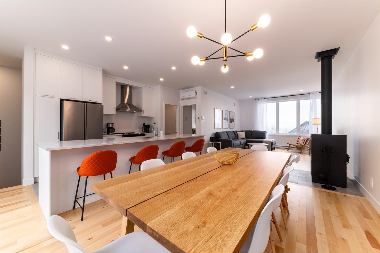 Cuisine moderne avec îlot, tabourets orange, et table en bois. Salle de séjour à l'arrière.