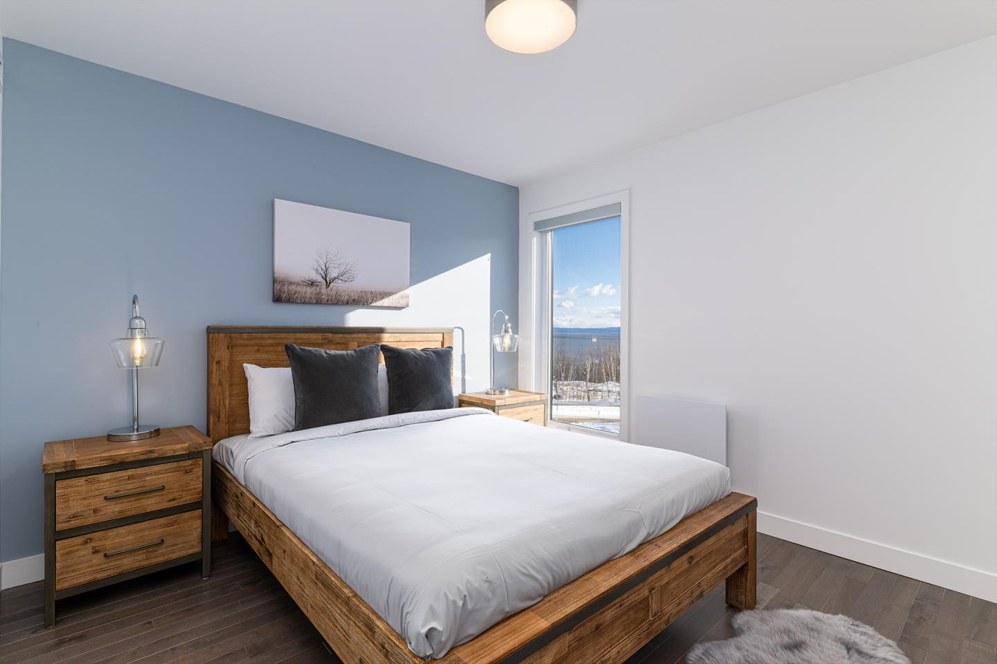 Chambre lumineuse avec lit en bois, mur bleu et grande fenêtre.