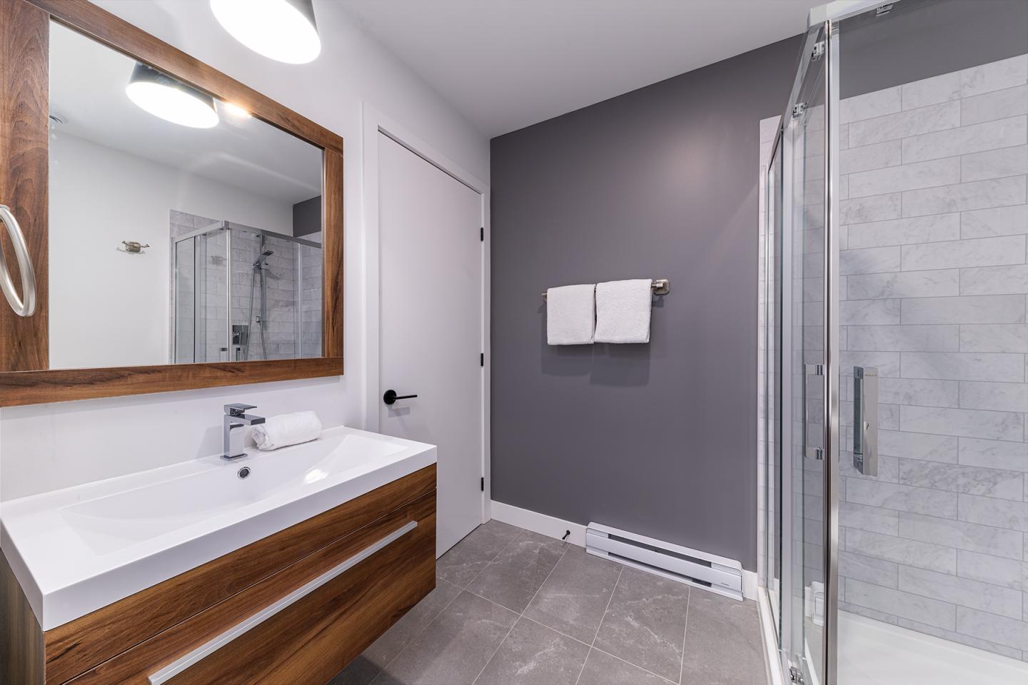 Salle de bain moderne, murs gris, douche vitrée, meuble bois avec lavabo blanc.