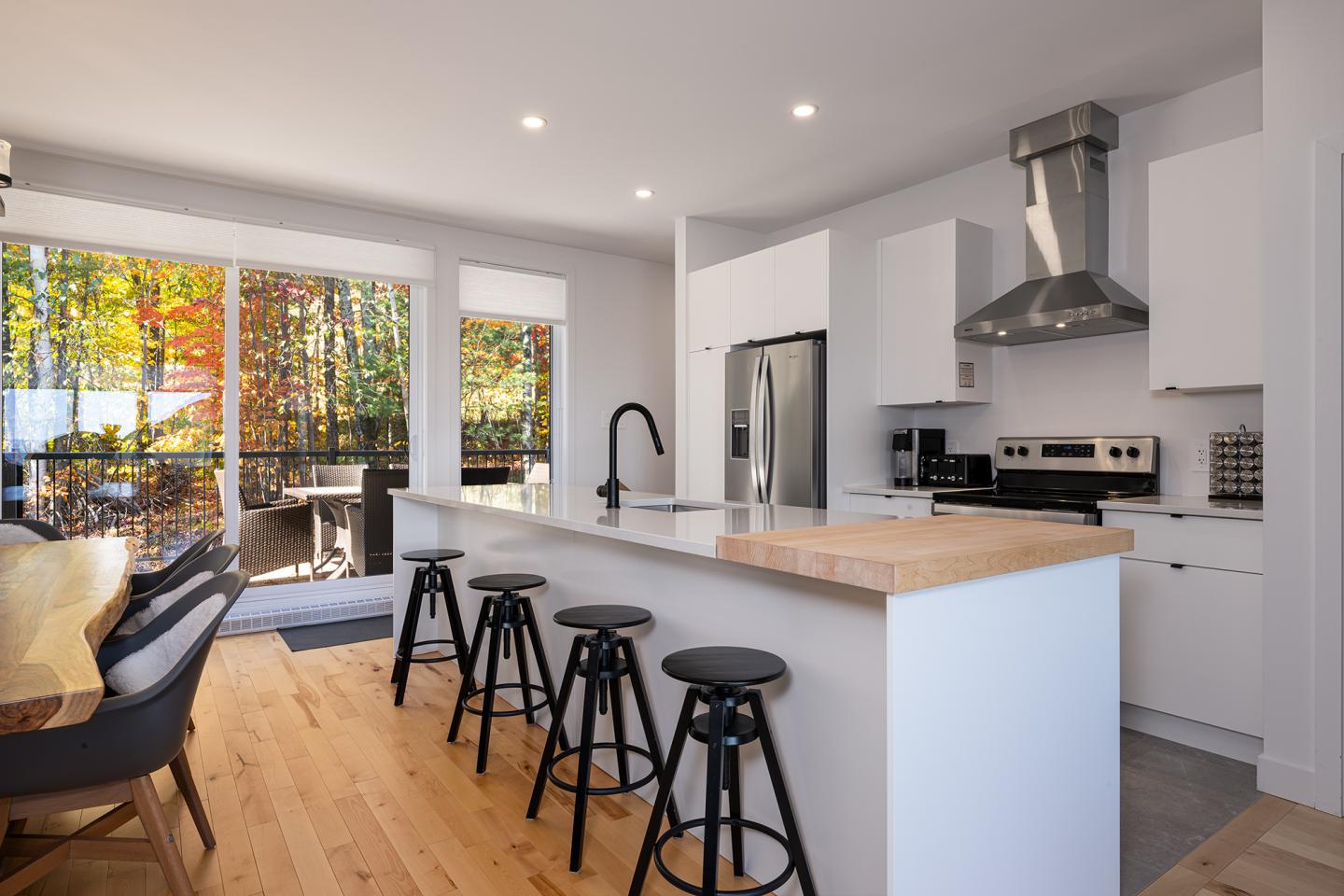 Cuisine moderne avec îlot central, tabourets noirs et baie vitrée donnant sur jardin.