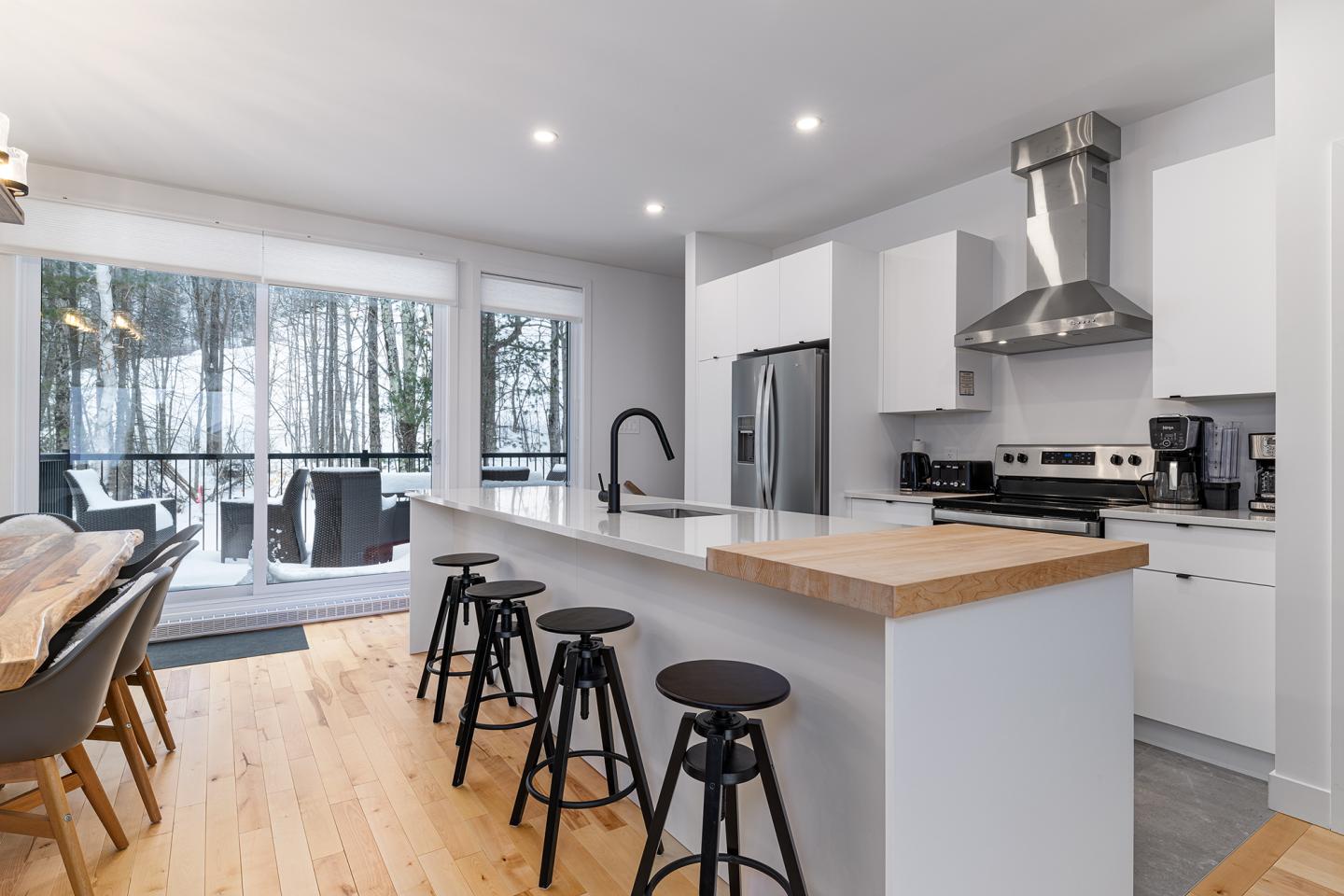 Cuisine moderne avec îlot, tabourets, et vue sur forêt enneigée à travers de grandes fenêtres.
